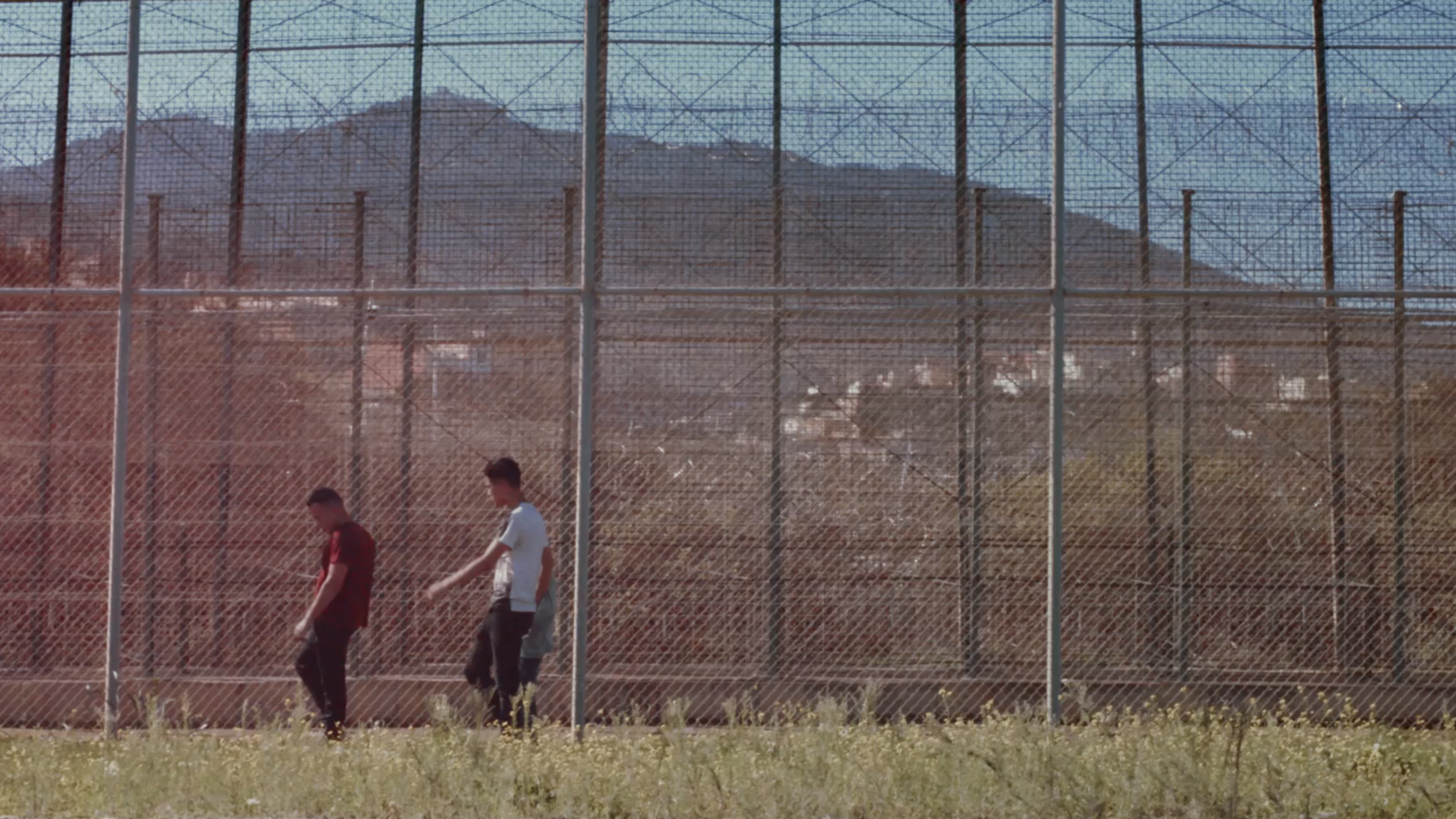 Dos jóvenes caminan junto a una valla perimetral en un entorno cerrado, imagen de un documental sobre menores no acompañados en Melilla. Remember my name