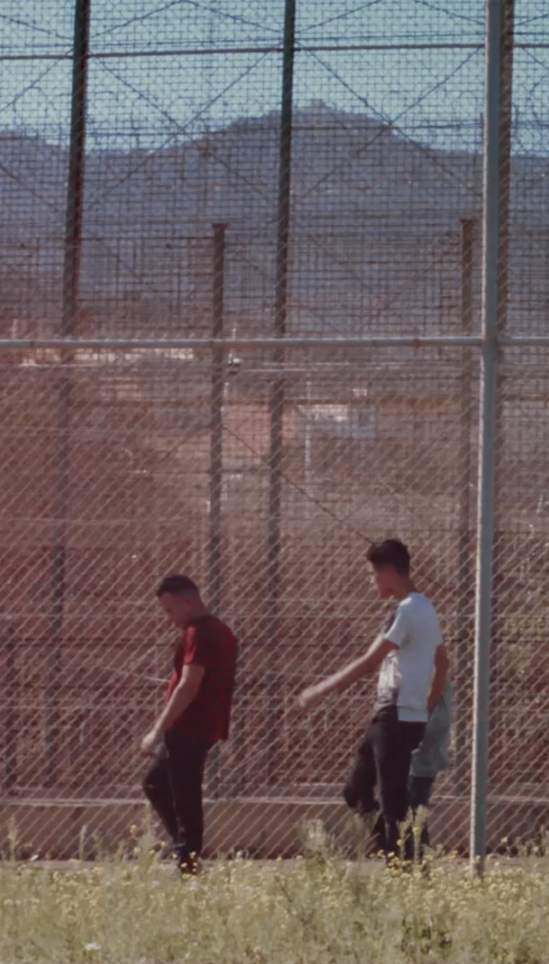 Dos jóvenes caminan junto a una valla perimetral en un entorno cerrado, imagen de un documental sobre menores no acompañados en Melilla. Remember my name